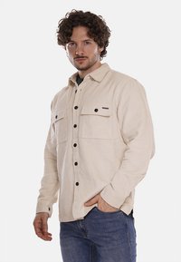 Camicia in cordonato beige con chiusura a bottoni, due tasche sul petto e maniche lunghe, realizzata con un tessuto testurizzato e bottoni scuri.