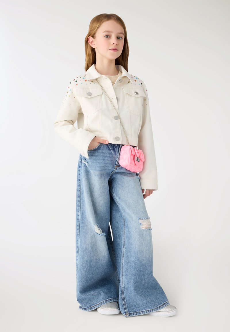 Młoda dziewczyna stoi z ręką w kieszeni, ubrana w białą kurtkę z kolorowymi ćwiekami, szerokie rozdarcia na dżinsach, srebrne buty i małą różową torebkę.