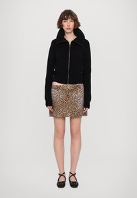 Svart stickad zip-up-tröja med hög krage, parat med en mini-skjorta i tan och svart leopardmönster. Modell som bär svarta flats med korsade remmar.