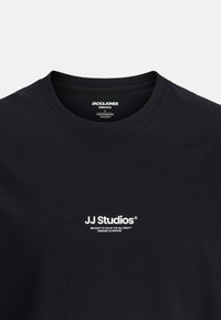 Schwarzes Baumwoll-T-Shirt mit Rundhalsausschnitt und kurzen Ärmeln. Mit weißem Textdruck "JJ Studios" auf der Brust. Etikett innen sichtbar.