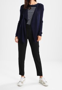 Cárdigan navy con diseño de frente abierto sobre una camiseta de rayas, combinado con pantalones negros y zapatillas blancas estampadas. Tejido suave, ajuste relajado.