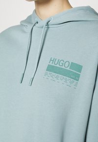 Sudadera de la marca Hugo en algodón azul claro, con un gráfico frontal en verde, cordón ajustable y un acabado suave y texturizado.