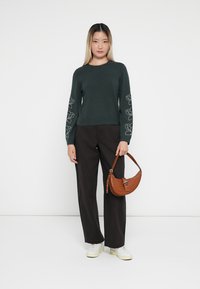 Pull vert foncé avec broderie florale sur les manches, associé à un pantalon noir à jambes larges et un sac à main en cuir marron. Des baskets blanches complètent le look.