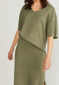 Olivgrön stickad klänning med korta ärmar, V-ringning och en sidoklyfta. Slät, texturerad tyg med avslappnad passform och draperad design.