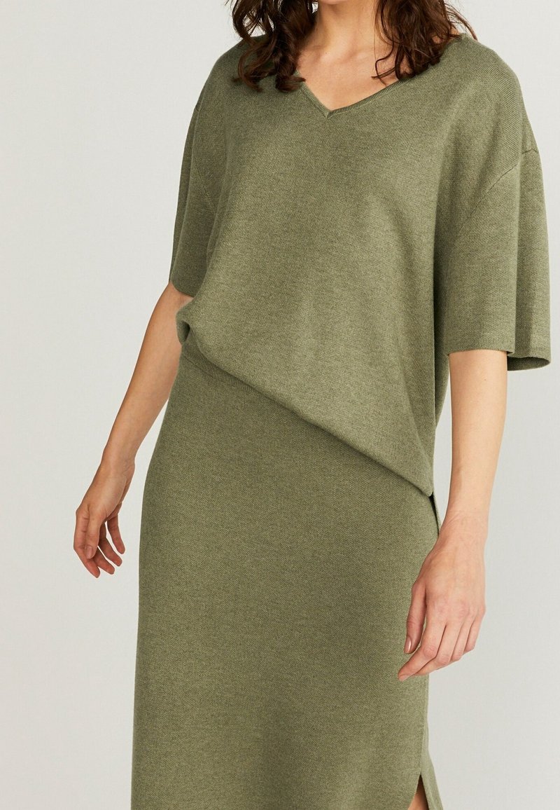 Olivgrön stickad klänning med korta ärmar, V-ringning och en sidoklyfta. Slät, texturerad tyg med avslappnad passform och draperad design.