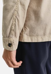 Beige Baumwolljacke mit sichtbaren Nähten, Knopfmanschetten-Details und strukturiertem Stoff; kombiniert mit dunklen marineblauen Hosen.
