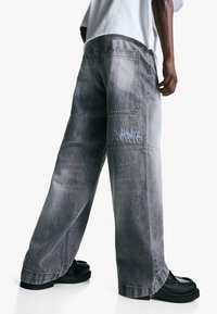 Spodnie dżinsowe w szerokim kroju, w wyblakłym szarym kolorze, z wieloma kieszeniami i haftem w stylu graffiti w jasnoniebieskim kolorze w pobliżu dołu. Teksturowany materiał, luźny krój.