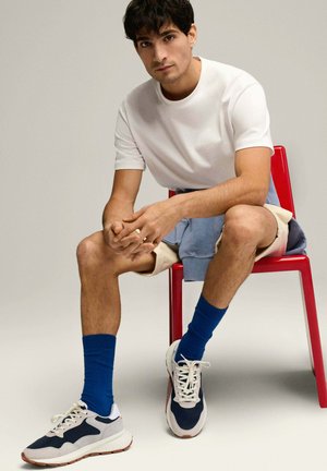 Camiseta blanca, pantalones cortos beige, calcetines azules y zapatillas deportivas en azul marino y blanco. Sentado en una silla roja. Fondo neutro.