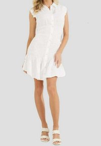 Weißes Knopfkleid mit kurzen Ärmeln, gerafftem Oberteil und gestuftem Volantsaum. Kombiniert mit weißen sandalen mit geflochtenen Riemen.