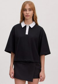 Zwarte oversized polo met korte mouwen en een witte kraag en knopenlijst. Gecombineerd met een zwarte wikkelrok. Soepele textuur.