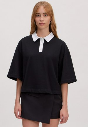 Zwarte oversized polo met korte mouwen en een witte kraag en knopenlijst. Gecombineerd met een zwarte wikkelrok. Soepele textuur.