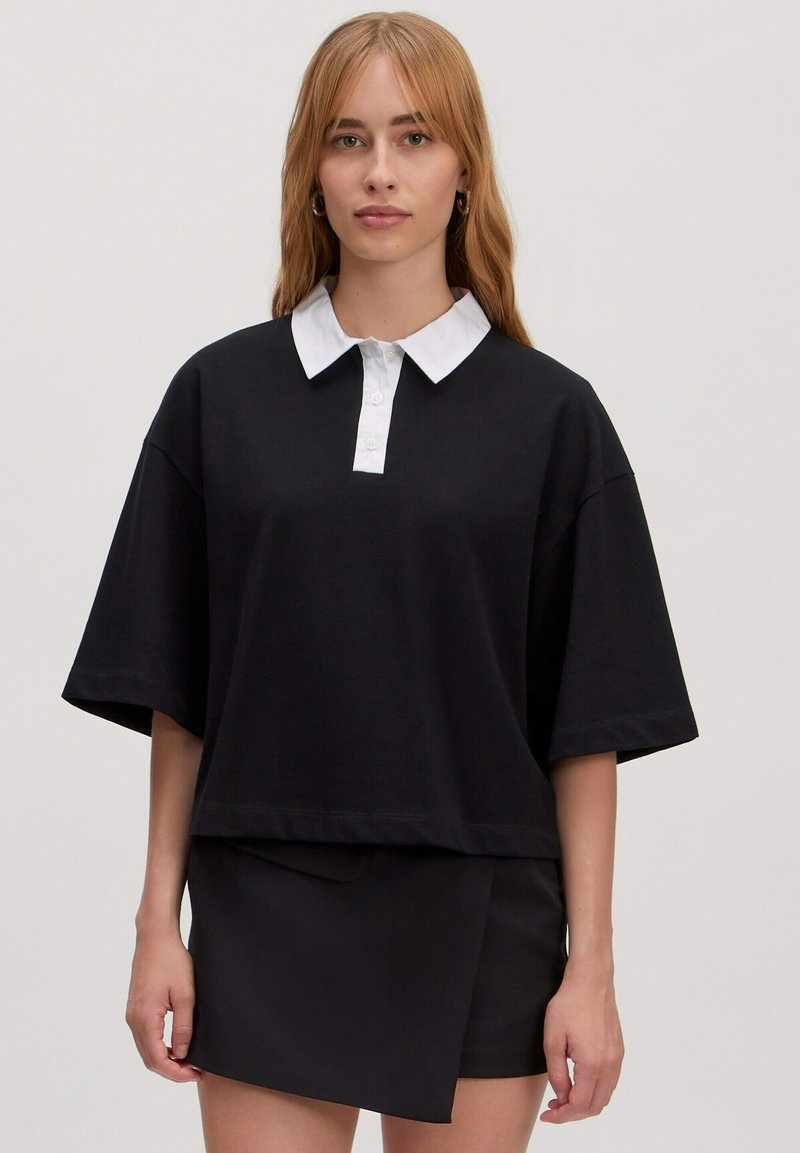 Zwarte oversized polo met korte mouwen en een witte kraag en knopenlijst. Gecombineerd met een zwarte wikkelrok. Soepele textuur.