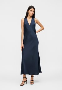 Γυναίκα που φοράει αμάνικο μακρύ φόρεμα σε σκούρο μπλε με V λαιμόκοψη και πλαϊνές σκισμές, ποζάροντας με το ένα χέρι στη μέση και φορόντας μαύρα πέδιλα με λουράκια.