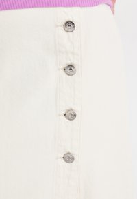 Jupe en denim blanc avec cinq accents de boutons en argent sur le côté, avec des coutures visibles et une surface de tissu légèrement texturée.