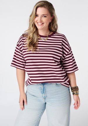 Lachende vrouw met golvend blond haar, draagend een bordeauxrood en roze gestreept hemd, lichtblauwe spijkerbroek, gouden sieraden en armbanden.
