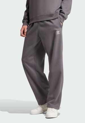 Pantalones de chándal de algodón gris con un ajuste holgado, que presentan un logo blanco en el muslo izquierdo y bolsillos laterales. Llevados con zapatillas deportivas blancas.