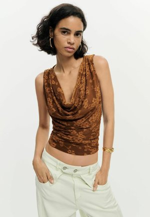 Młoda kobieta o krótkich ciemnych włosach, ubrana w bezrękawnik w brązowy kwiatowy wzór i białe spodnie, z rękami w kieszeniach, pozująca na białym tle.