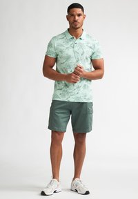 Vaaleanvihreä poolopaita, jossa on kasviaiheinen kuvio, yhdistetty oliivinvihreisiin cargo-shortseihin. Malli käyttää valkoisia urheilukenkiä, joissa on harmaat yksityiskohdat.