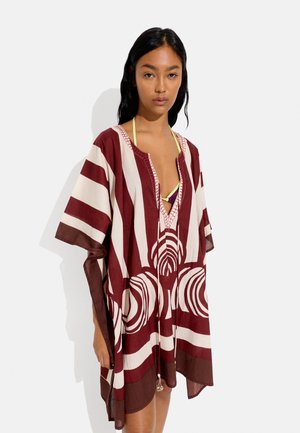 Kobieta ubrana w luźny kaftan w bordowo-białe paski z okrągłymi wzorami na żółtym topie bikini, stojąca na jednolitym tle.
