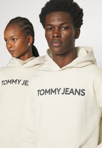 Dos individuos llevan sudaderas beige con "TOMMY JEANS" impreso en letras negras y en negrita. La tela parece suave y tiene un corte relajado; las capuchas están forradas.