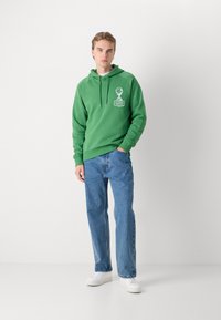 Πράσινη φούτερ με μπροστινή γραφική εκτύπωση, κουκούλα με κορδόνια, ριμπ μανσέτες, συνδυασμένη με μπλε τζιν και λευκά αθλητικά παπούτσια. Απλός σχεδιασμός, χαλαρή εφαρμογή.