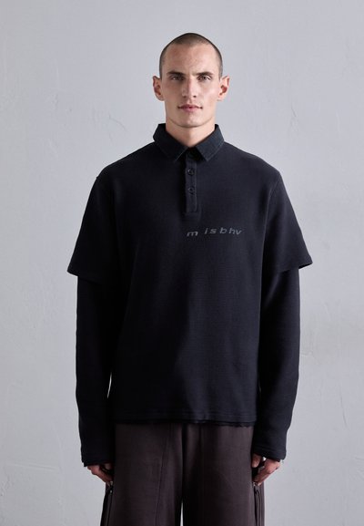 Mann mit kurzem Haar, der ein mehrschichtiges schwarzes, strukturiertes Langarmshirt mit Polokragen trägt und vor einer schlichten hellgrauen Wand steht.
