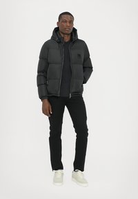Schwarze Pufferjacke mit Kapuze, Reißverschluss vorne und Seitentaschen. Getragen über einem schwarzen Shirt und kombiniert mit schwarzen Hosen und hellen Turnschuhen.