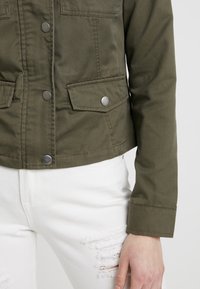 Olivově zelená zkrácená bunda vyrobená z odolného materiálu, se zapínáním na patentky a dvěma předními kapsami, stylizovaná s bílými opotřebovanými džíny.