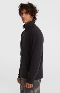 Giacca in pile nera con zip, colletto alto e maniche lunghe, realizzata in materiale morbido; indossata con pantaloni stampati camouflage.