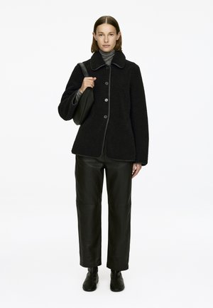 Schwarze Fleecejacke mit einem Kragen, Knopfverschluss und kontrastierenden Details. Kombiniert mit schwarzen Lederhosen und Stiefeletten.