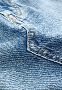 Nahaufnahme von Denimstoff mit einem hellblauen Ton und sichtbarer Struktur, gelben Kontrastnähten und einem Detail der Taschenkante.