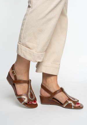 Feet wearing brown wedge sandals with metallic accents and beige rolled-up pants against a plain background.