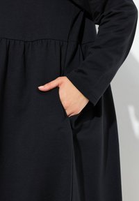 Schwarzes langärmliges Kleid aus weichem Stoff, mit gerafftem Taillenbund und einer seitlichen Tasche, glatten Nähten und ohne sichtbare Muster.