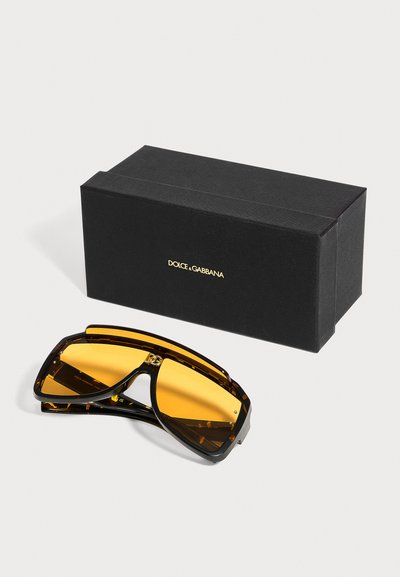 Μαύρα γυαλιά ηλίου Dolce & Gabbana με μεγάλα κεχριμπαρένια φακούς τοποθετημένα δίπλα σε μια μαύρη θήκη με χρυσό λογότυπο σε λευκό φόντο.