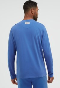 Mann in blauem Langarmhemd und Hose, von hinten gesehen, mit einem weißen Etikett nahe dem Nacken mit der Aufschrift „No#1 focus on colour CAMP DAVID.“