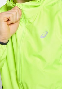 Veste légère verte brillante avec fermeture éclair, capuche et logo réfléchissant. Équipée de poignets élastiques et d'un tissu lisse et texturé.