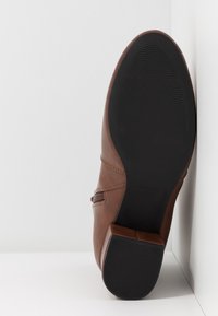 Botte en cuir marron avec une semelle noire texturée et un petit talon bloc, positionnée verticalement contre un fond blanc en coin.