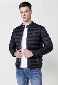 Chaqueta acolchada negra con acolchado horizontal, cierre de cremallera y cuello alzado. Presenta un pequeño logo en el lado izquierdo del pecho y una cremallera en contraste.