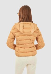 Chaqueta puffer color arena con un diseño ajustado, acolchado horizontal y capucha. Presenta una textura lisa y brillante, así como puños elásticos. Vista trasera.