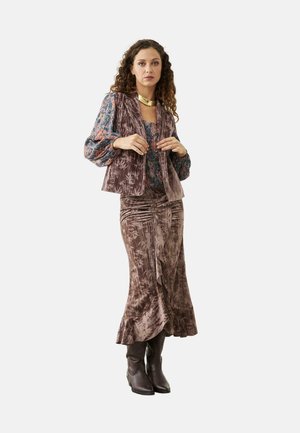 Mujer con cabello rizado que lleva un chaleco y falda de terciopelo morado con estampado, blusa floral, collar dorado y botas marrón oscuro.