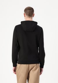 Hanorac negru cu glugă, cu mâneci lungi, textură fină, design simplu și manșete cu riburi. Modelul poartă pantaloni bej.