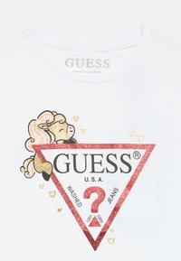 Biała koszulka z dużym logo "GUESS" w czerwono-złotym brokatowym kolorze w kształcie trójkąta. Zawiera zabawną grafikę z kucykiem i drobnymi serduszkami.