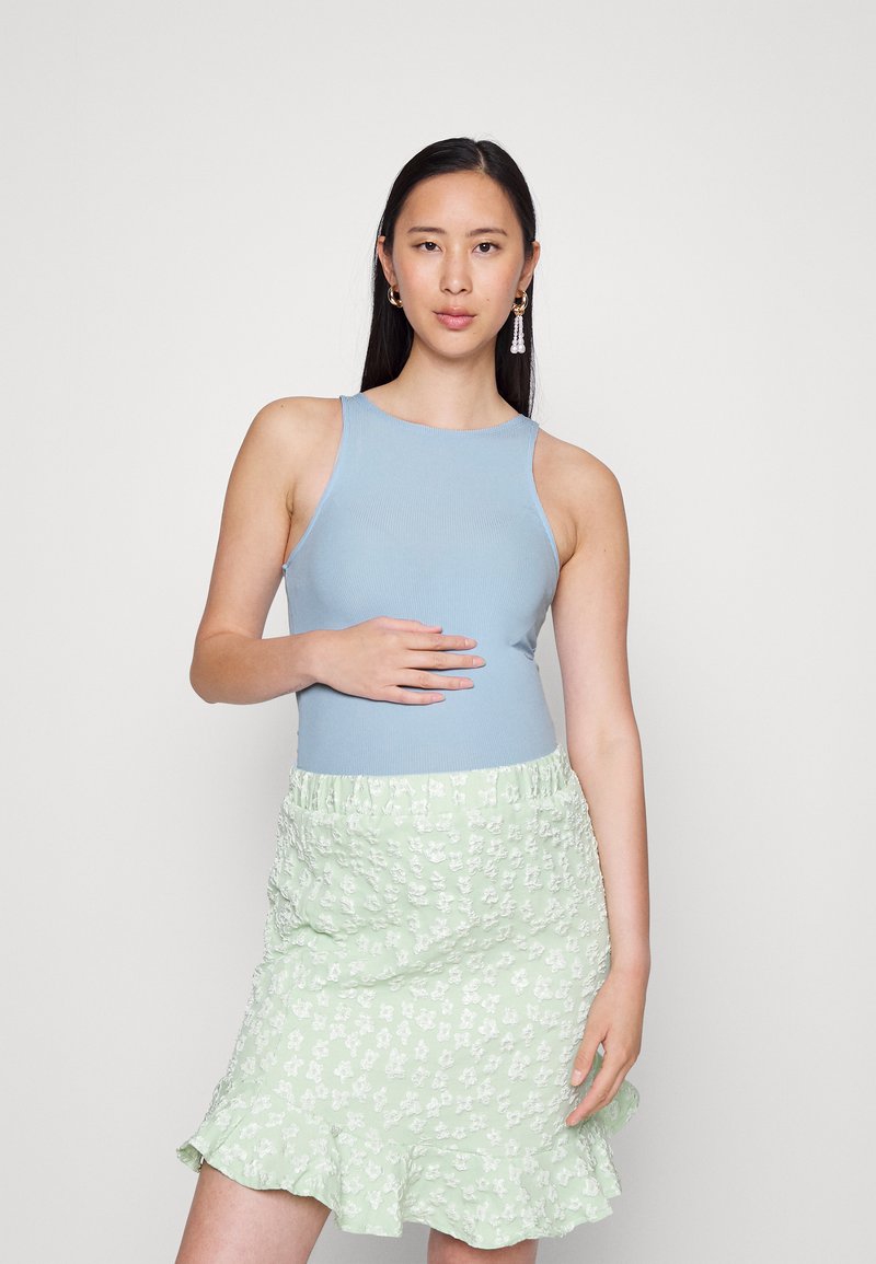 Top de tanque ribete azul claro combinado com uma saia verde menta com babados, apresentando pequenos padrões florais brancos. O modelo está posando em um fundo neutro.
