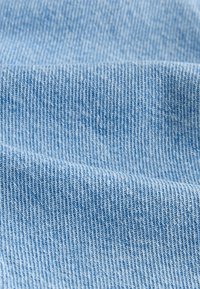 Denimstof in lichtblauw, met een fijne geribbelde textuur en zichtbare stiksels die een patroon van diagonale lijnen vormen.