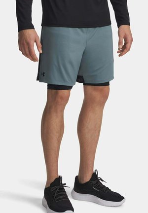 Mann trägt graue Sportshorts mit schwarzen Kompressionsshorts darunter und schwarzen Laufschuhen auf weißem Hintergrund.