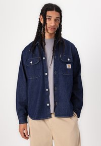Denim shirt in dark blue with two chest pockets, snap buttons, and contrasting stitching. Worn over a light gray T-shirt.