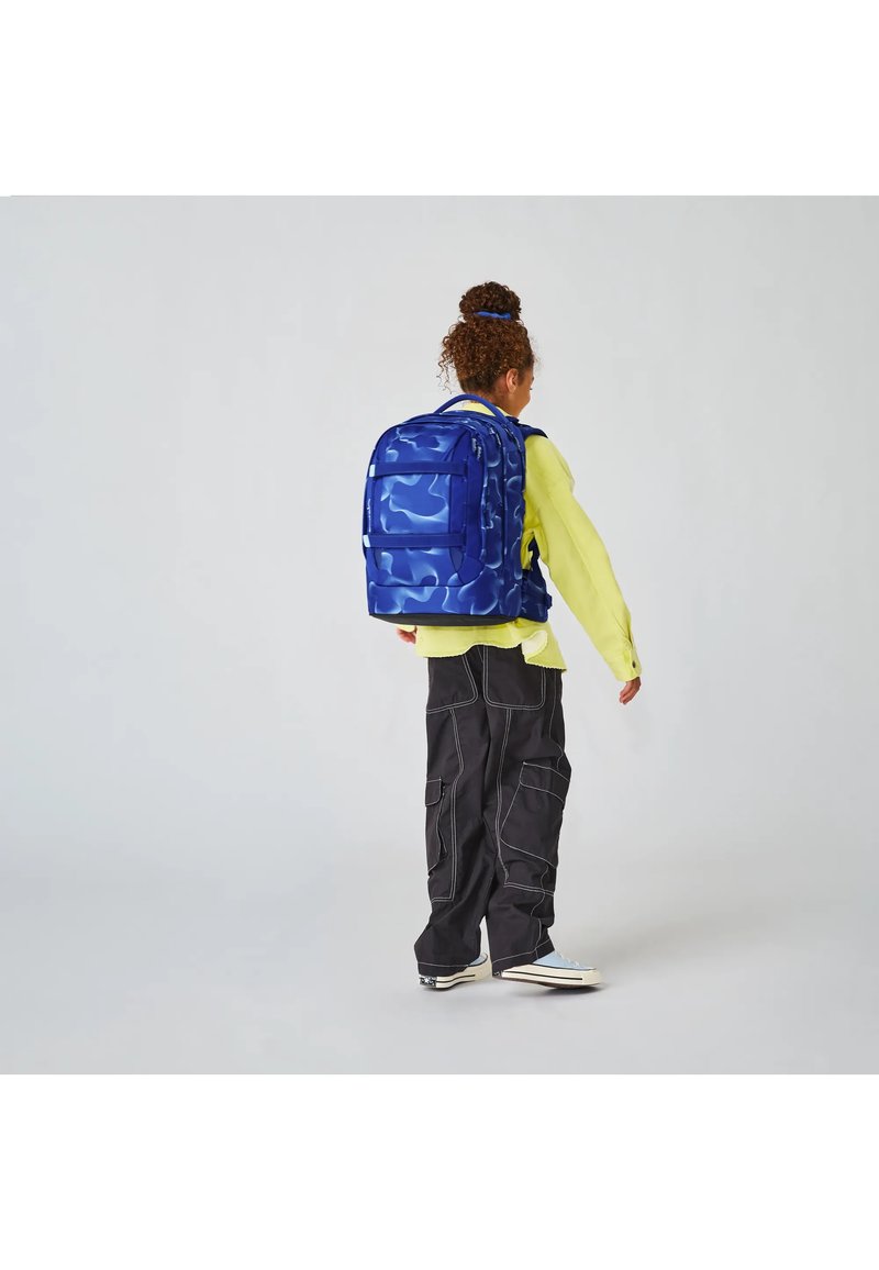 Kind mit lockigem Haar im Dutt trägt einen leuchtend gelben Pullover, schwarze Cargo-Hosen und einen großen blauen Rucksack mit Tarnmuster, steht seitlich.