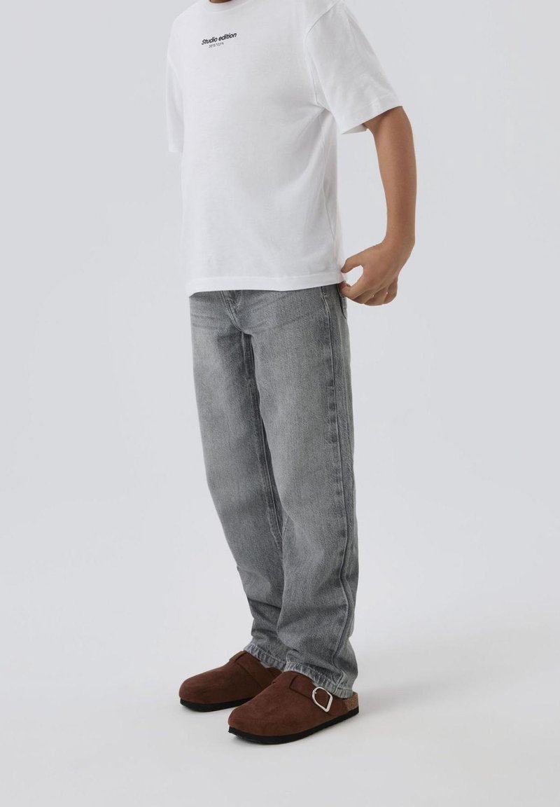 Personne portant un t-shirt blanc à manches courtes, un jean gris et des chaussures marron sans lacets avec un détail de boucle, debout devant un fond clair uni.