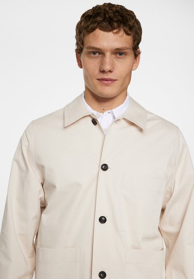 Licht beige jack met een kraag, zwarte knopen, een voorzak en een soepele, matte textuur.