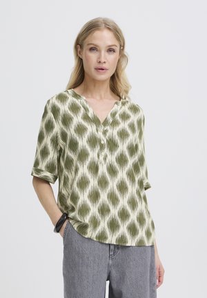 Femme portant un chemisier à motif vert olive avec des boutons et des manches retroussées, associé à un pantalon gris et un bracelet noir, debout devant un fond blanc.